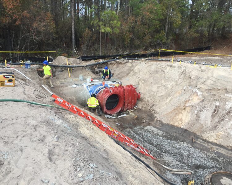 Workers install red and blue pipeline in trench, trees nearby.