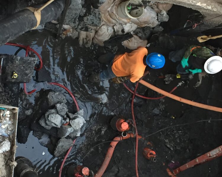 Two construction workers in safety gear work in a muddy, confined space.