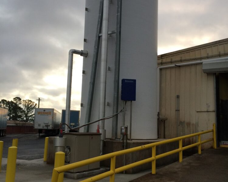Tall industrial tank with pipes near beige building, yellow railings.