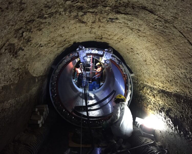 Workers in protective gear work on a large pipe underground.