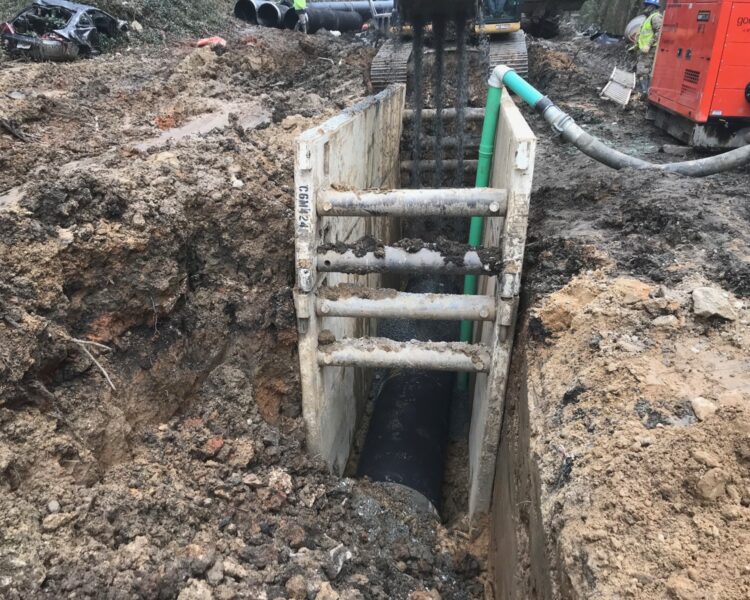 Zona de excavación con cajón de zanja, tubería a la vista y maquinaria.