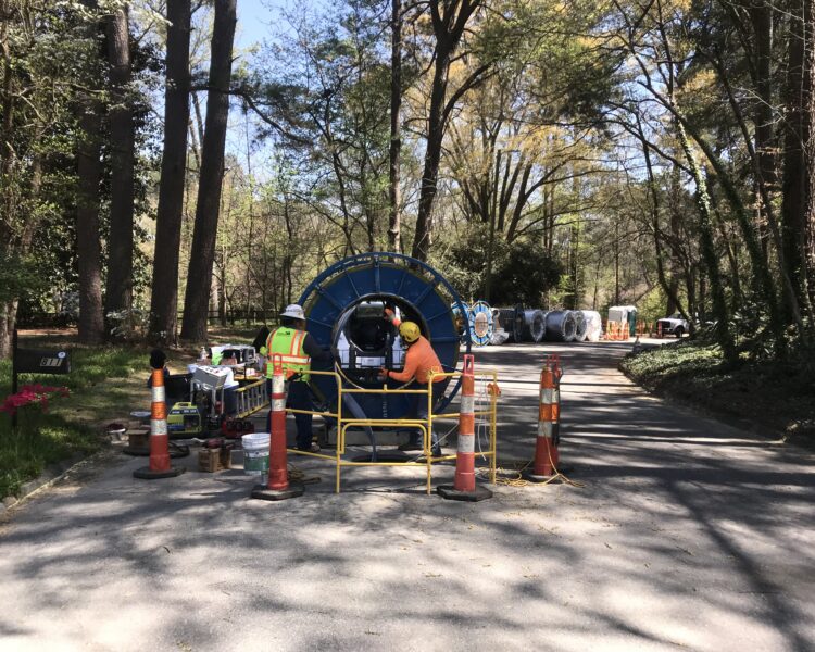 Two construction workers use equipment on a street with cones.