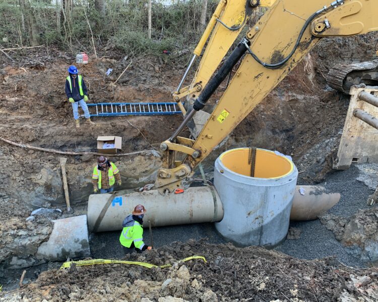 Workers guide a concrete pipe moved by excavator outdoors.