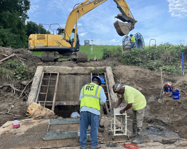 Los trabajadores instalan equipos junto a una estructura de drenaje; una excavadora trabaja en las inmediaciones.