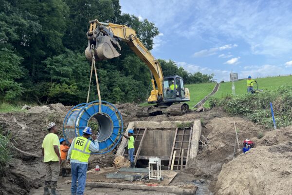 Un equipo levanta un tubo azul con una excavadora en una obra rural sin asfaltar.