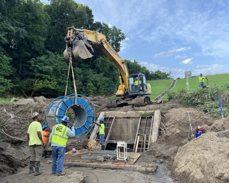 Un equipo levanta un tubo azul con una excavadora en una obra rural sin asfaltar.