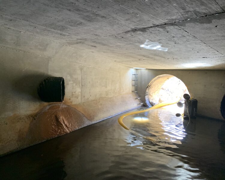 Túnel de hormigón, agua estancada, manguera amarilla, escalera hacia una salida iluminada por el sol.