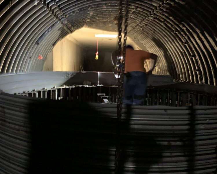 Un trabajador con una camiseta naranja está de pie en un túnel en penumbra con tuberías.