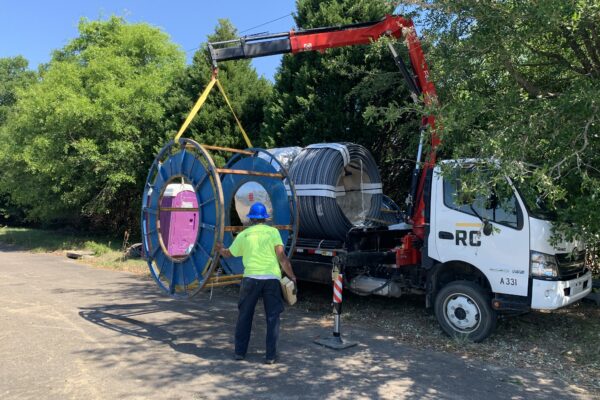 Un trabajador guía una bobina izada por una grúa con un camión teledirigido entre los árboles.