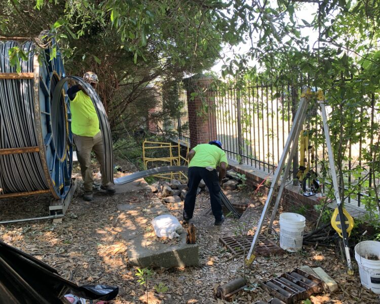 Unos trabajadores, equipados con material de seguridad, manipulan unos grandes cables al aire libre cerca de una valla.