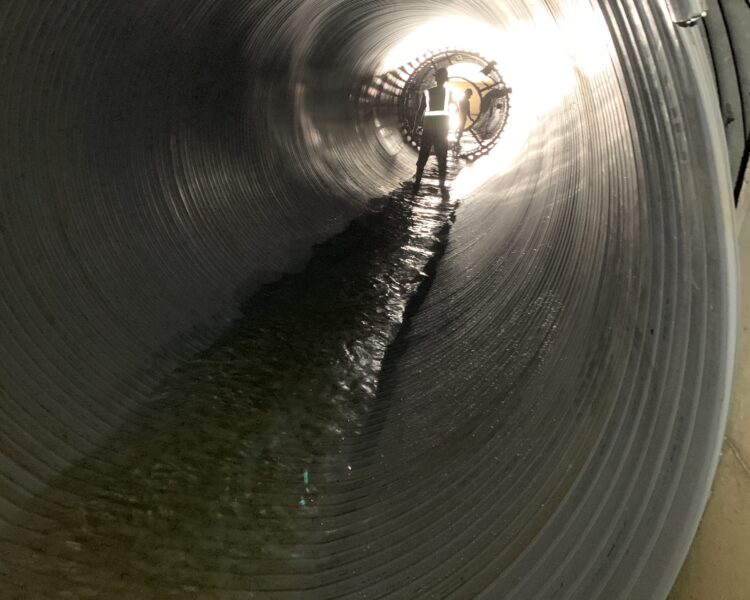 Un trabajador en un túnel de gran tamaño con agua poco profunda y luces intensas al frente.
