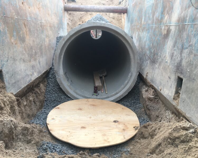 Concrete pipe partly buried in gravel, tools and plywood nearby.
