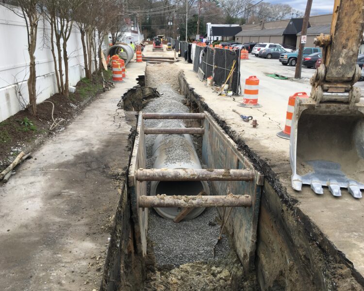Pipe installation in trench with gravel, excavator, cones, barriers.