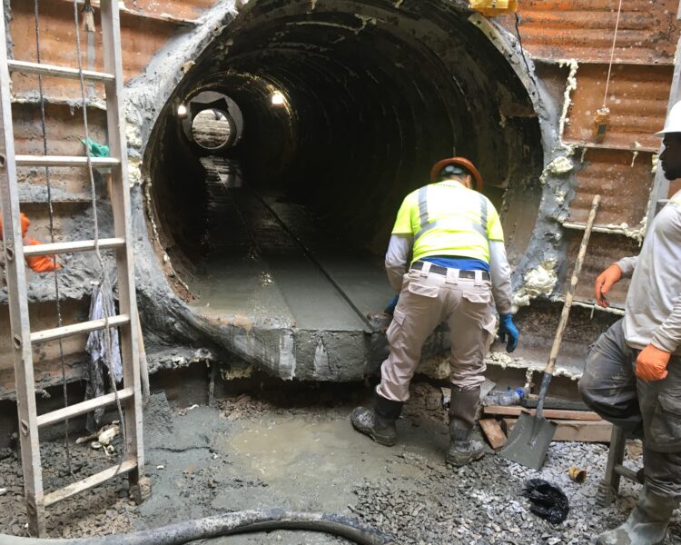 Two construction workers in safety gear at concrete tunnel entrance.