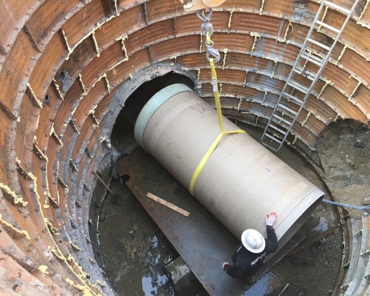 Worker in hard hat guides concrete pipe into deep shaft.