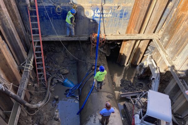 Unos operarios equipados con material de seguridad reparan una gran tubería en un foso lleno de barro.