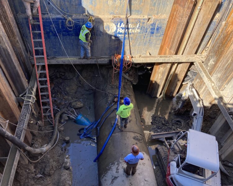 Unos operarios equipados con material de seguridad reparan una gran tubería en un foso lleno de barro.