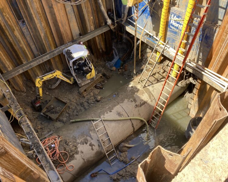 Zanja en una obra con tuberías, excavadora, escaleras, mangueras y maquinaria.