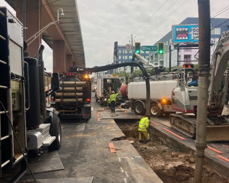 Unos trabajadores y varios vehículos reparan la carretera bajo las vías del tren elevado.