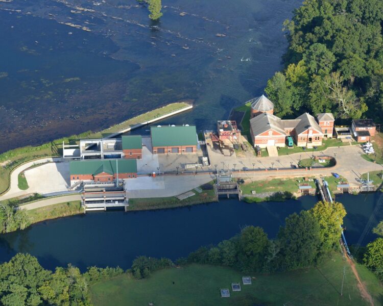 Aerial shot of riverside facility, canal, dam, and trees.