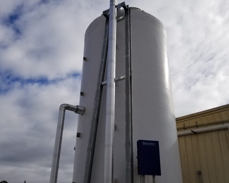 White industrial tank with vertical pipes by a yellow building.