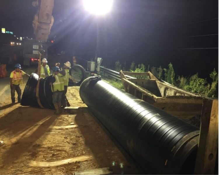 Workers in safety gear install a large pipe at night.