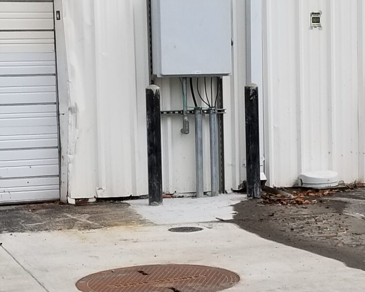 Gray electrical box with red button, two black posts, manhole.