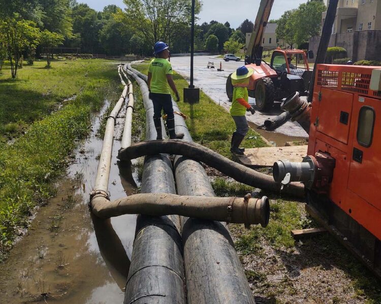 Workers in safety gear set up black hoses by orange machine.