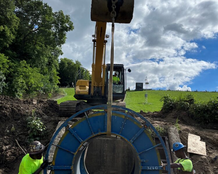 Los trabajadores guían un gran anillo metálico que una excavadora está levantando al aire libre.