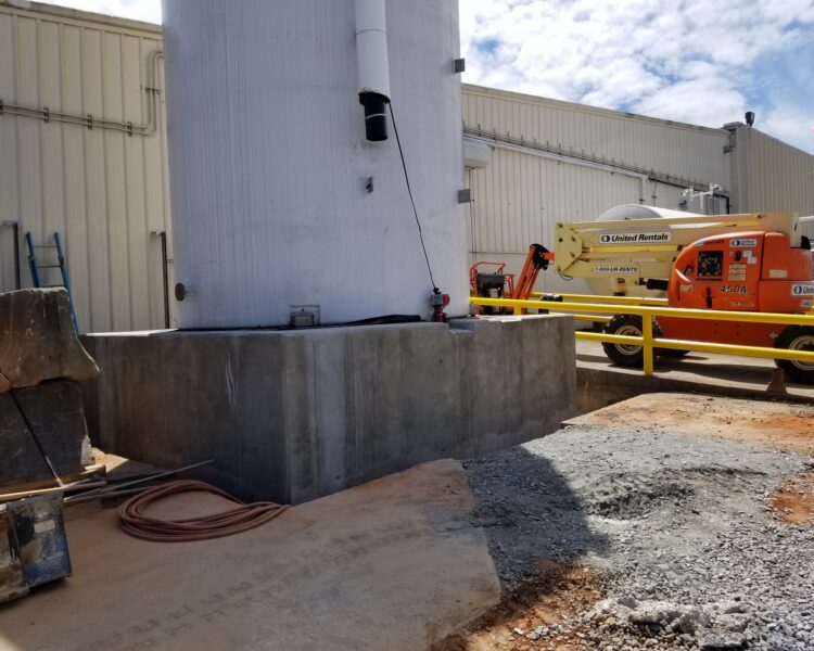 Large outdoor tank on concrete base beside beige building and lift.