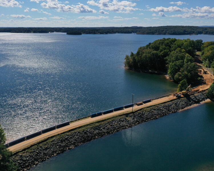 Train crosses narrow causeway between water and trees, aerial view.