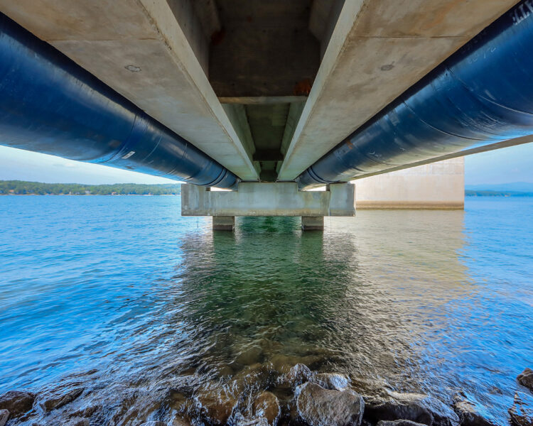 Under a concrete bridge with blue pipes over clear water.