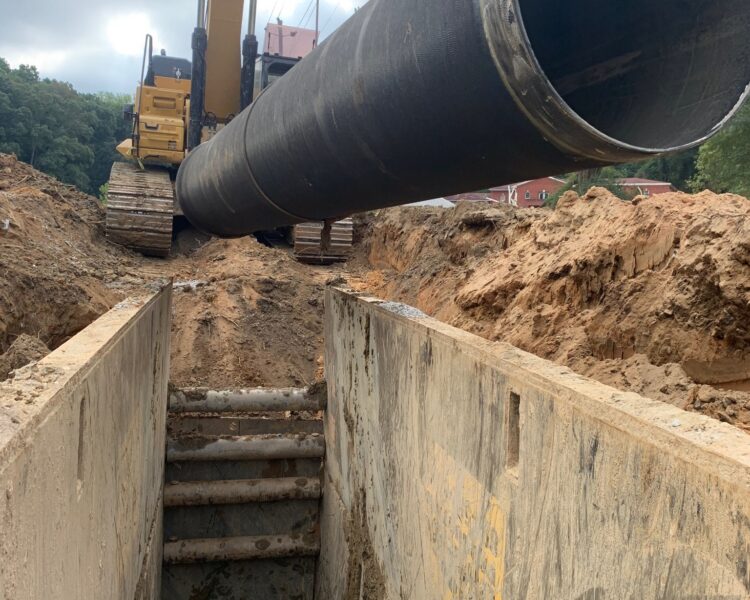 Una excavadora levanta un tubo de acero sobre una zanja embarrada.