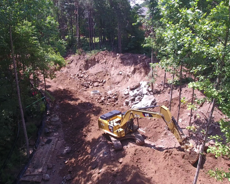 Yellow excavator digging beside trees and piles of soil.