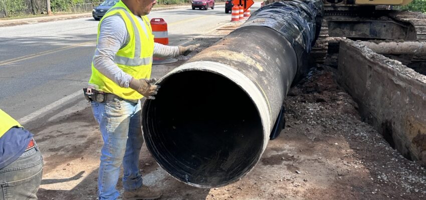 Workers guide a pipe moved by excavator near a road.