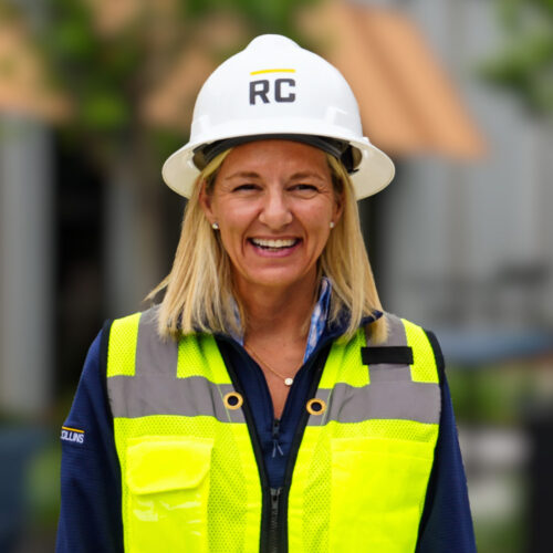 Smiling woman in RC hard hat and yellow vest at construction site.