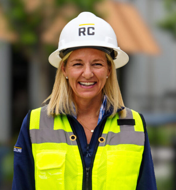 Smiling woman in RC hard hat and yellow vest at construction site.